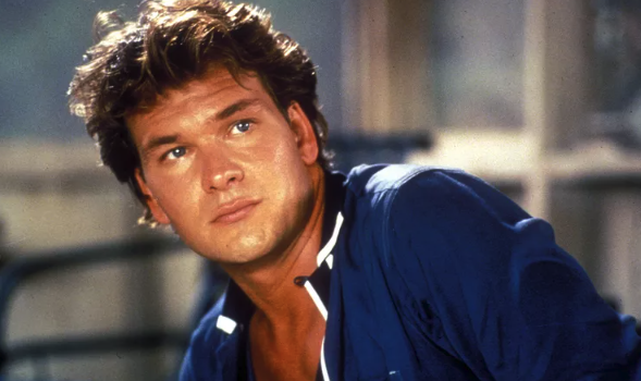 Patrick Swayze as Johnny Castle, a light-skinned man with short, styled brown hair wearing a casual blue button-down hair is looking off camera.